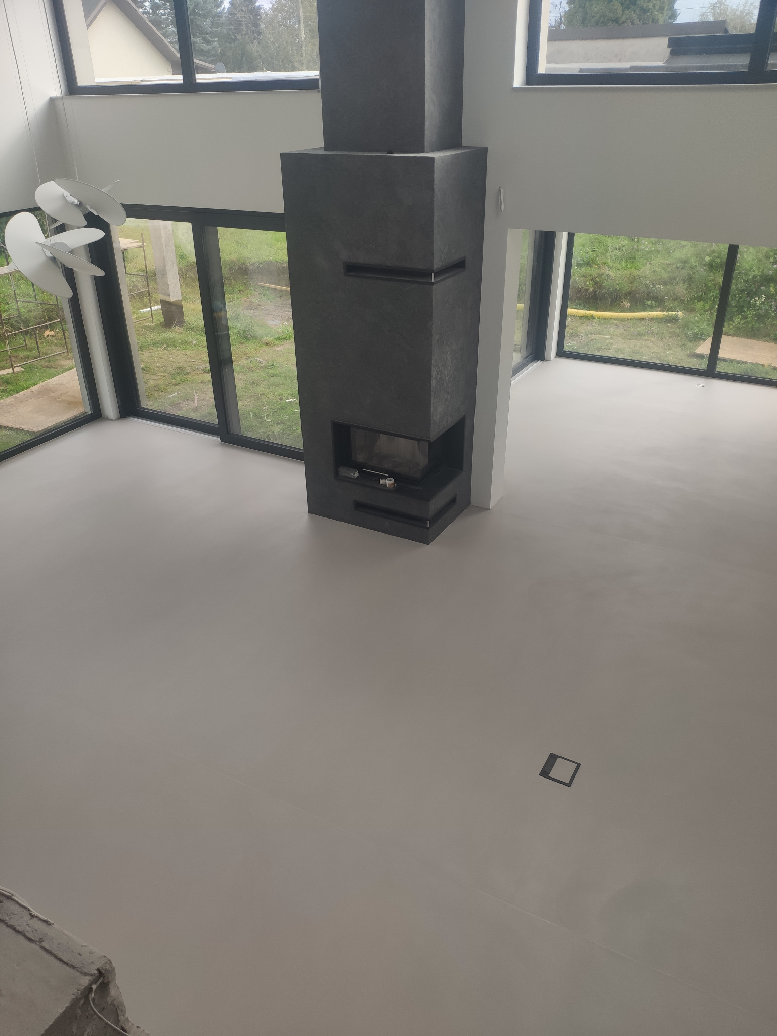 Jasnoszara posadzka z mikrocementu w nowoczesnym salonie z kominkiem i dużymi oknami. Widok z góry na gładką powierzchnię i minimalistyczny design.