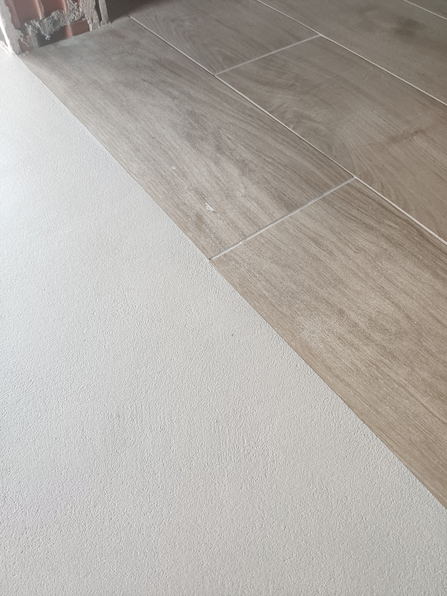 Połączenie gładkiego, jasnego mikrocementu na ścianie z drewnopodobną, ułożoną pod kątem podłogą z płytek. Widoczny fragment surowej ściany z cegły.