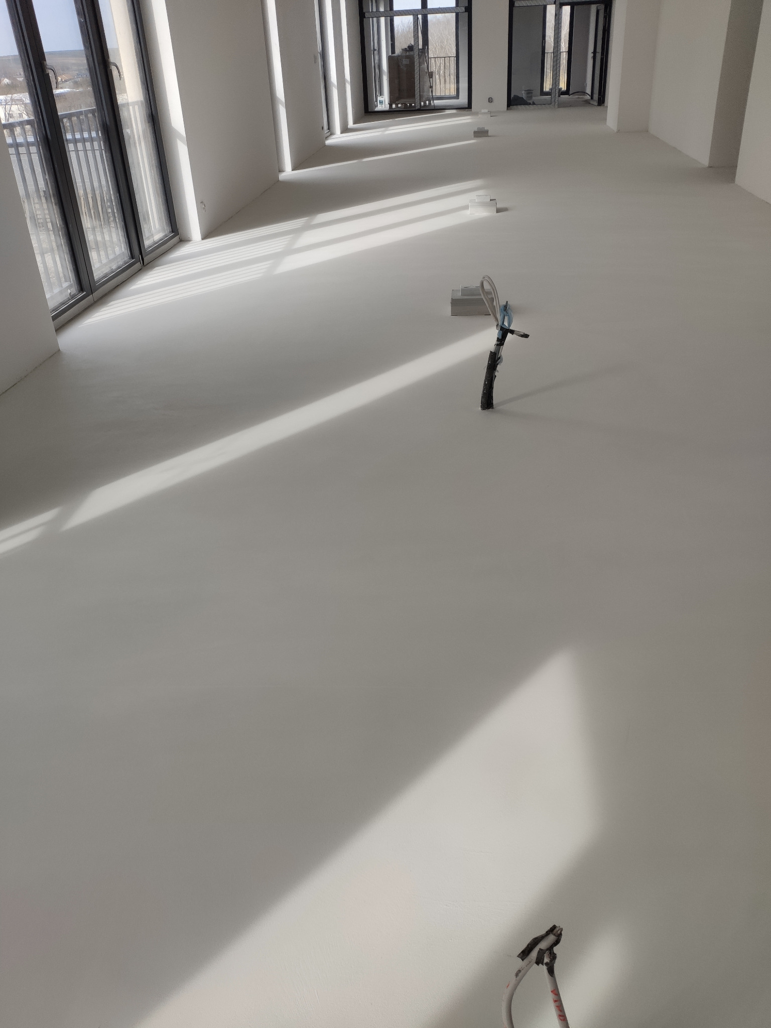 Jasna, gładka posadzka w przestronnym, nowoczesnym wnętrzu z dużymi oknami. Widoczne narzędzia i kable sugerują etap wykończeniowy. Minimalistyczny design, dominująca biel.