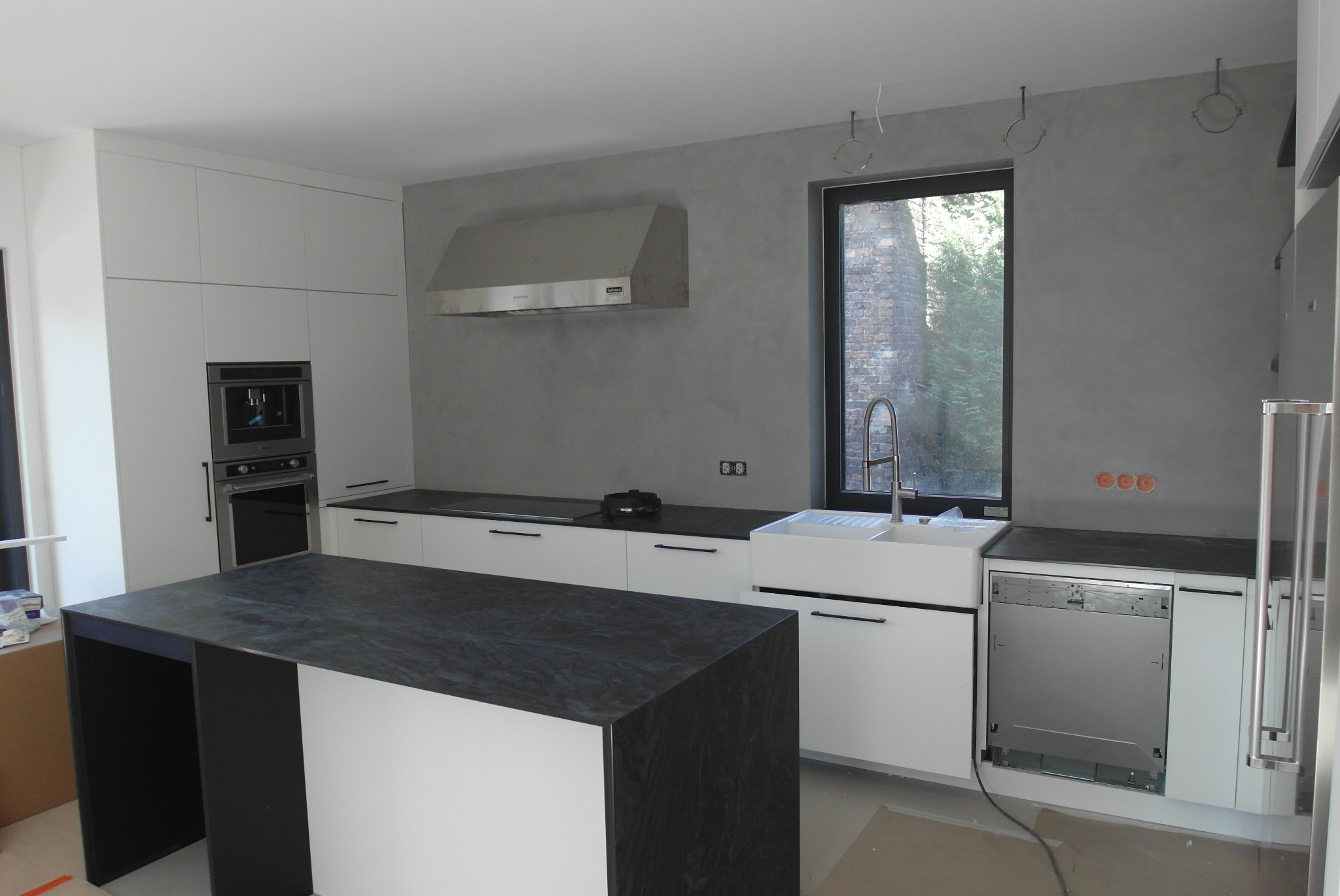 Nowoczesna kuchnia w trakcie wykończenia: białe szafki, czarny blat wyspy, ściana pokryta szarym mikrocementem. Widoczne niedokończone detale instalacji.