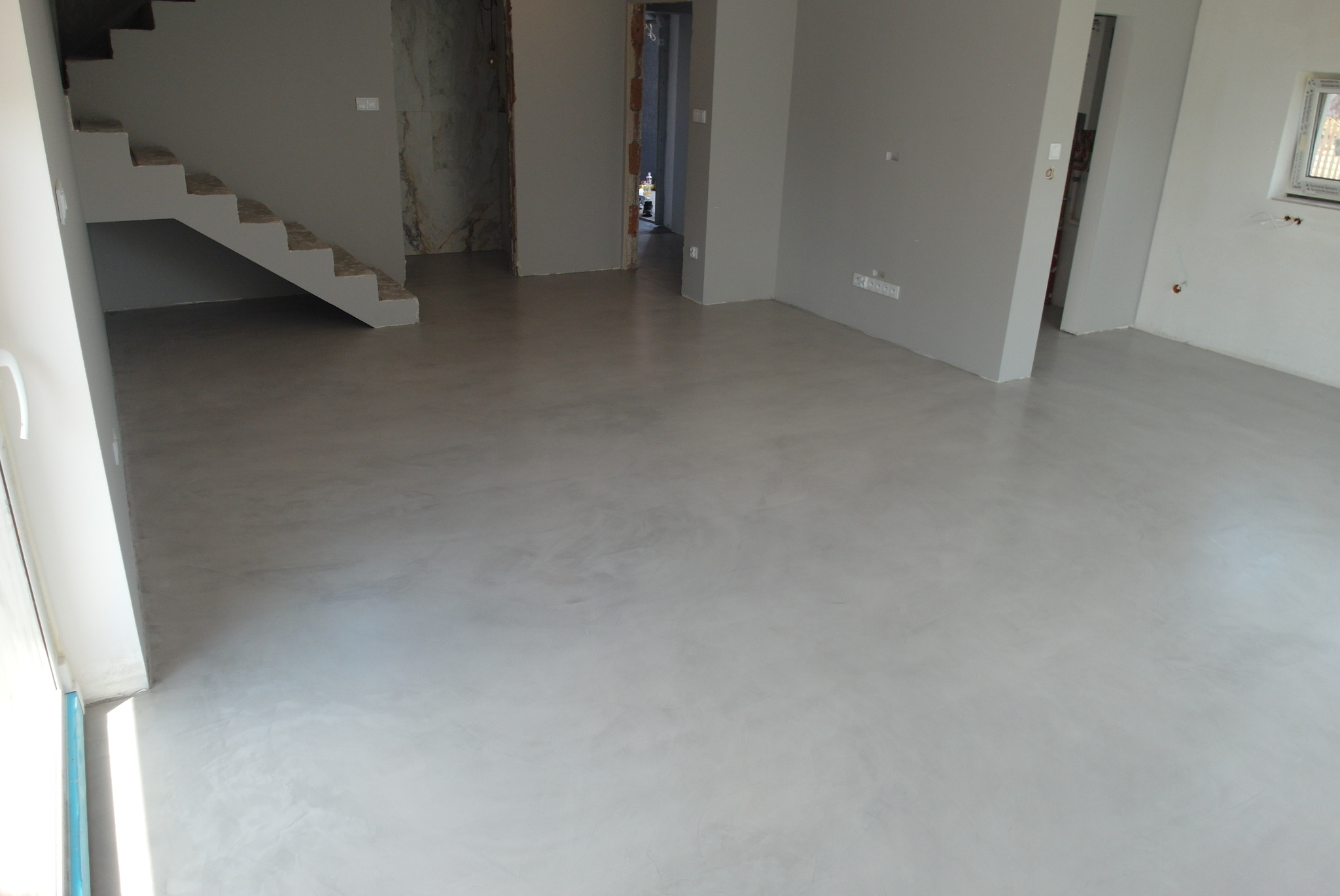 Jasnoszara, gładka posadzka z mikrocementu w przestronnym wnętrzu. Widoczne schody i surowe ściany, podkreślające minimalistyczny charakter wykończenia.