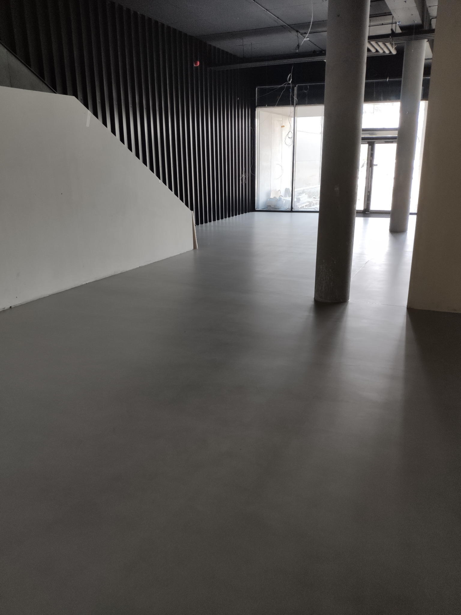 Szara, gładka posadzka w przestronnym wnętrzu z kolumnami i schodami. Minimalistyczny design, duże okna w tle. Widoczne elementy instalacji pod sufitem.