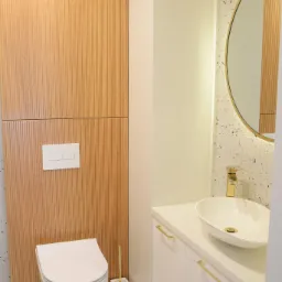 Nowoczesna łazienka z wiszącą toaletą na tle drewnianej ściany panelowej, okrągłym lustrem z podświetleniem LED i białą szafką z umywalką nablatową i złotymi uchwytami.