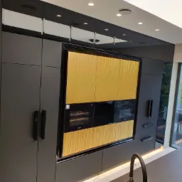 Nowoczesna zabudowa kuchenna w odcieniach szarości z elementami forniru w kolorze złota, wbudowane urządzenia AGD marki Miele, widok na taras przez duże okno.