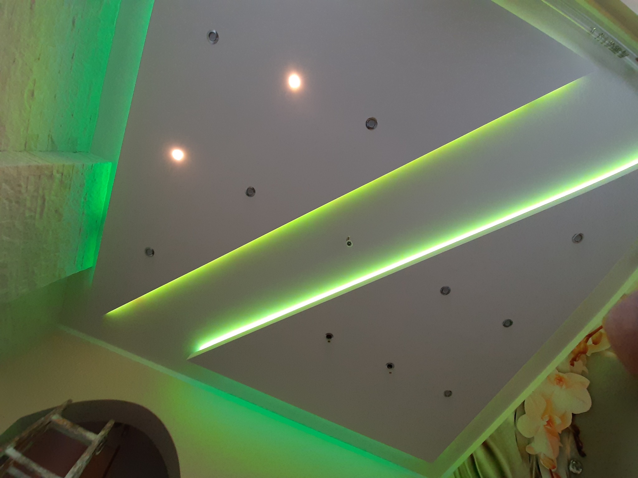 Nowoczesny sufit podwieszany z oświetleniem LED w odcieniach zieleni, widoczne halogeny, fragment ściany z teksturą i drabina w rogu pomieszczenia.