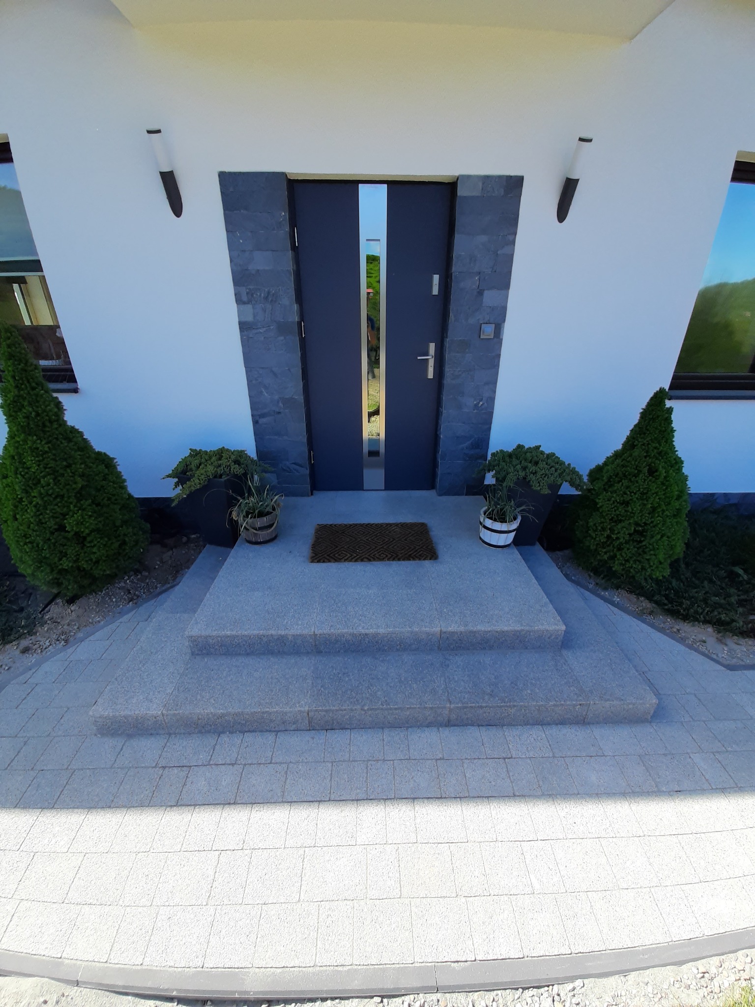 Nowoczesne wejście do domu z szarymi drzwiami, kamienną okładziną i trzema stopniami z kostki brukowej. Po bokach drzwi symetrycznie ustawione tuje i rośliny w donicach. Ściany białe z nowoczesnymi...