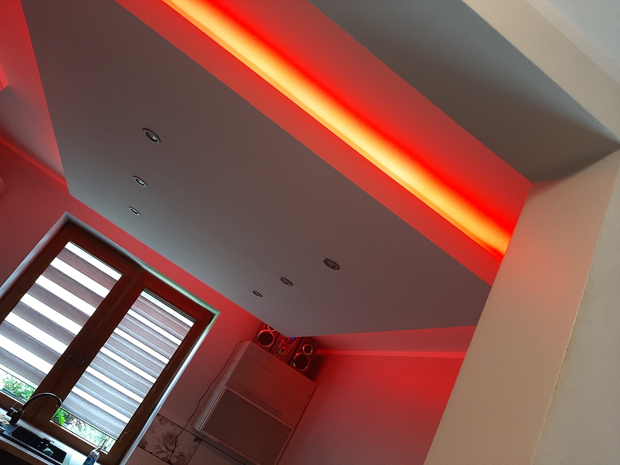 Nowoczesny sufit podwieszany z oświetleniem LED w kolorze czerwonym, widoczne okno z roletami dzień-noc i fragment zabudowy meblowej z głośnikami.