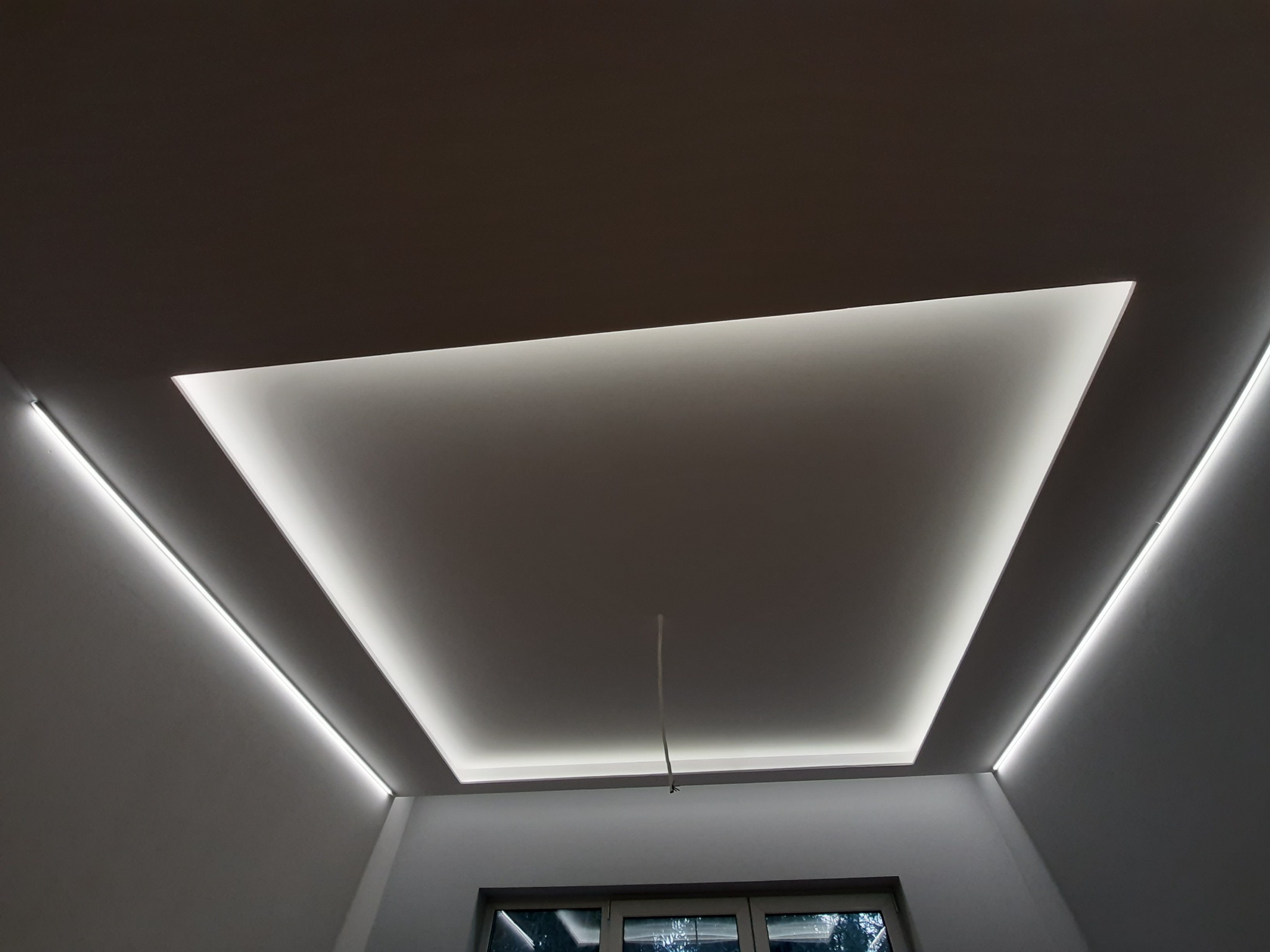 Nowoczesny sufit podwieszany z oświetleniem LED w kształcie prostokąta, z dodatkowymi liniami świetlnymi na ścianach, widok z dołu na okno.