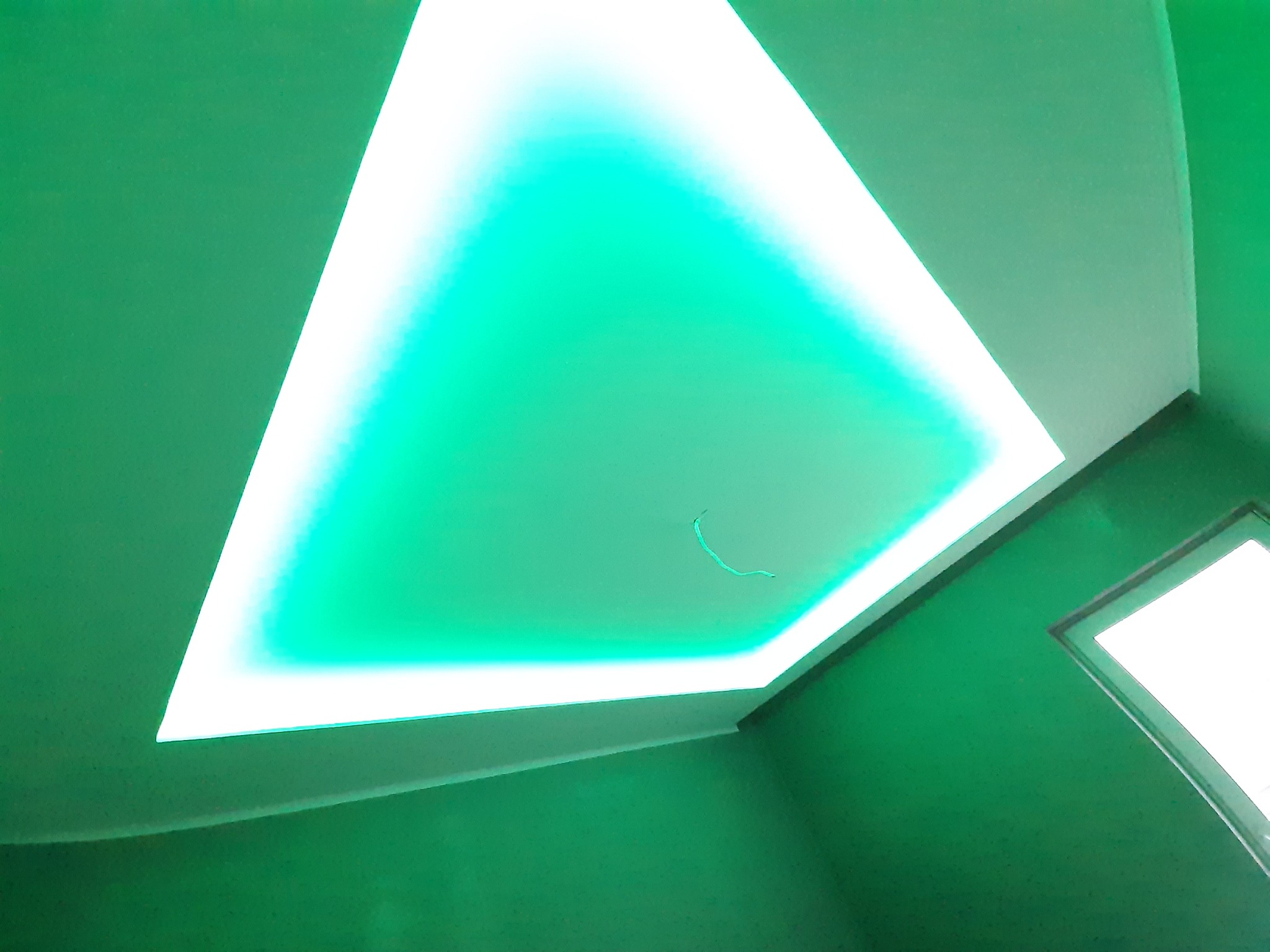 Nowoczesne oświetlenie sufitowe w kształcie trójkąta, emitujące intensywne, zielone światło, widoczne z perspektywy narożnika pomieszczenia z oknem w tle.