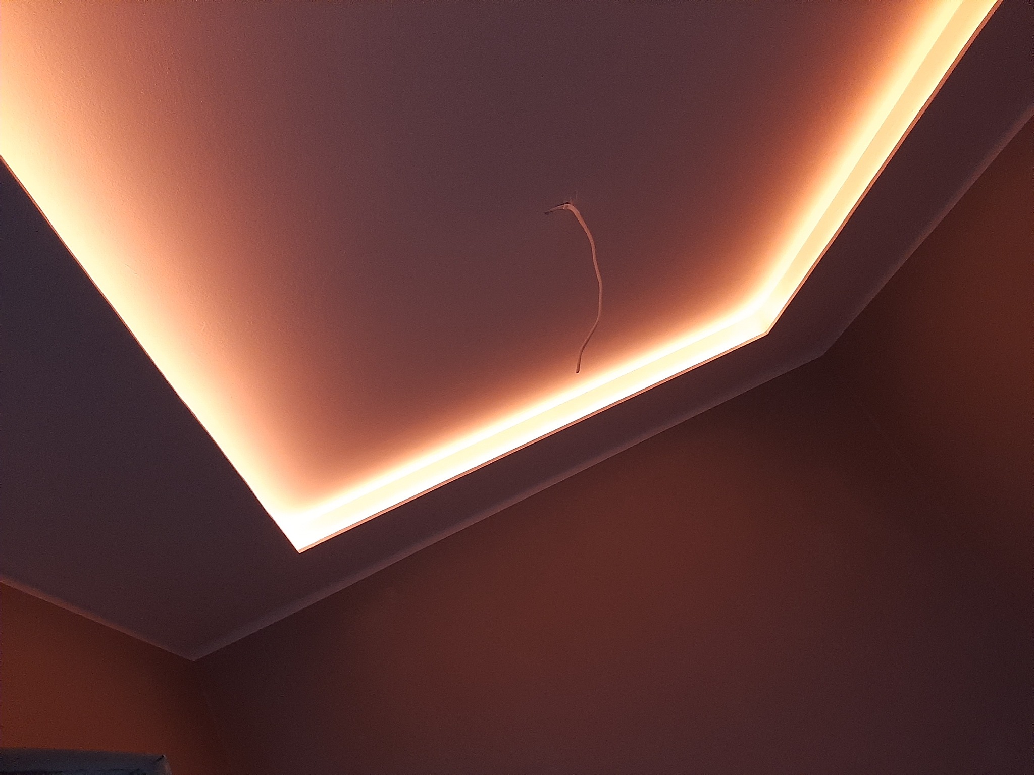 Sufit podwieszany z oświetleniem LED w ciepłej tonacji, widoczny wystający kabel elektryczny, ciemna ściana w tle.