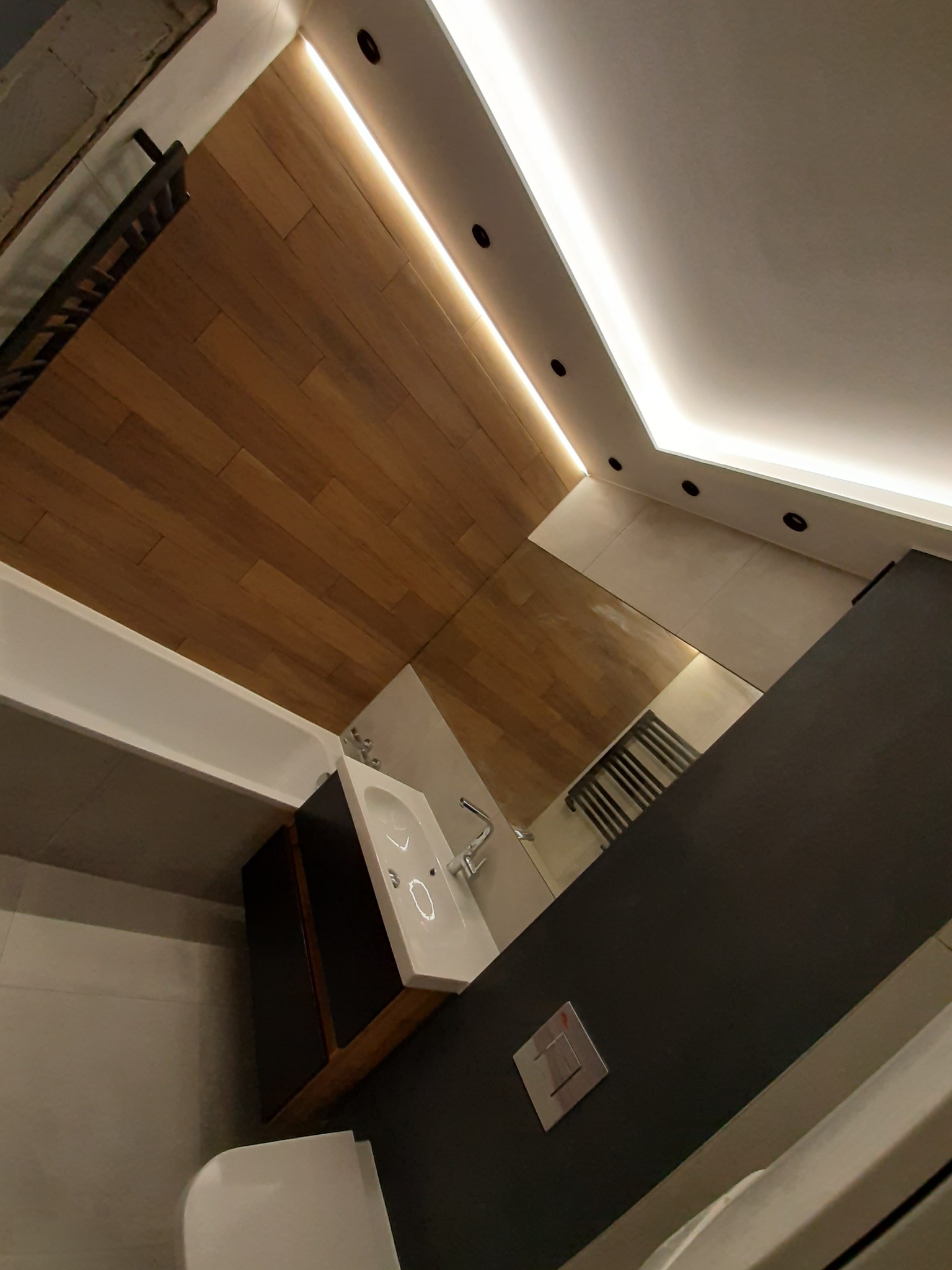 Nowoczesna łazienka z drewnianą okładziną sufitową, oświetleniem LED, umywalką nablatową na szafce z ciemnego drewna i grzejnikiem drabinkowym, sfotografowana pod nietypowym kątem, ukazując...