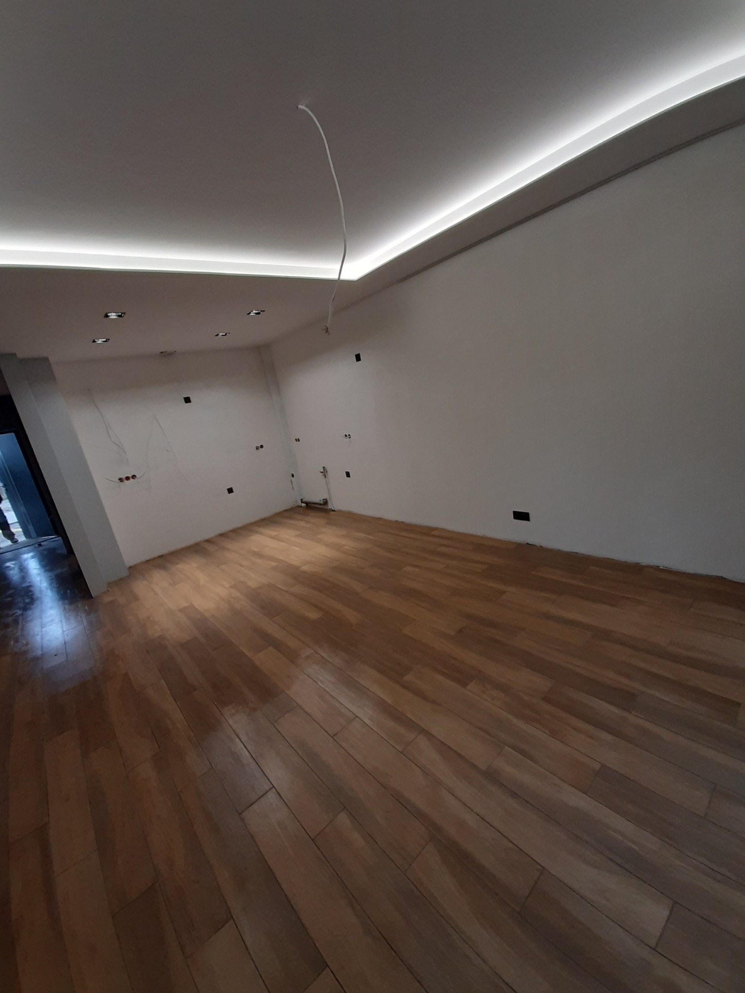 Pusty pokój po remoncie z drewnianą podłogą, białymi ścianami z widocznymi puszkami elektrycznymi i wystającymi kablami, oraz z podwieszanym sufitem z oświetleniem LED.