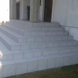 Zewnętrzne schody wejściowe do domu, wyłożone jasnoszarą, drobną mozaiką ceramiczną, z ciemnymi fugami, prowadzące do zadaszonego wejścia z kolumnami.