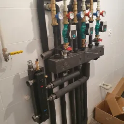 Rozdzielacz ogrzewania podłogowego z rurami w izolacji, manometrami i pompami obiegowymi na tle białych płytek, widoczne kartony na podłodze.