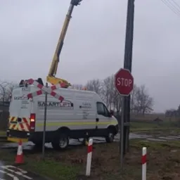 Pracownik w pomarańczowej kurtce w koszu podnośnika Smartlift naprawia linię energetyczną przy słupie z znakiem STOP, na tle pochmurnego nieba i wilgotnej drogi.