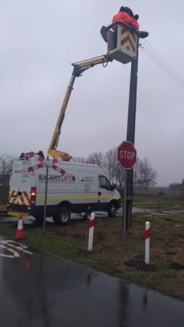 Pracownik w pomarańczowej kurtce w koszu podnośnika Smartlift naprawia linię energetyczną przy słupie z znakiem STOP, na tle pochmurnego nieba i wilgotnej drogi.