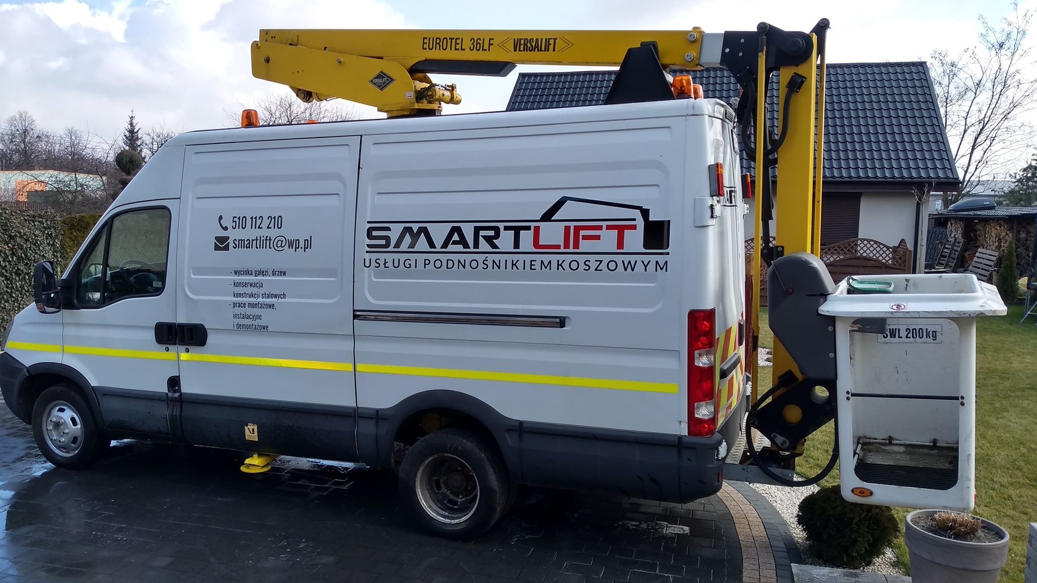 Biała furgonetka z żółtym wysięgnikiem koszowym zaparkowana na posesji, z widocznym logo firmy Smartlift i danymi kontaktowymi na boku.