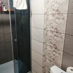 Wykończona łazienka z kabiną prysznicową z ciemnymi płytkami, biały ręcznik na kabinie, ściana wyłożona płytkami w odcieniach beżu z motywem kwiatowym, widoczna toaleta z rolką papieru toaletowego.