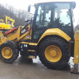 Żółta koparko-ładowarka Caterpillar 432F2 na mokrym placu budowy, w tle inne maszyny budowlane i drzewa bez liści.