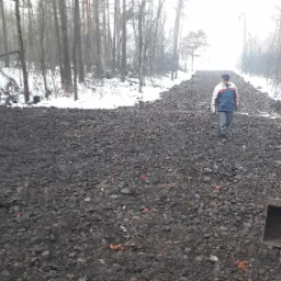 Widok z kabiny maszyny budowlanej na utwardzoną, kamienistą drogę przez zasypany śniegiem las; w oddali idzie mężczyzna w kurtce.