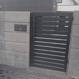 Nowoczesna, grafitowa brama wjazdowa z poziomymi szczeblami, zamontowana na betonowych słupkach z szarą skrzynką na listy z kodem, na tle domu z szarą elewacją i dachem.