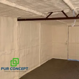 Wnętrze garażu po natrysku pianką PUR, widoczne ściany, sufit i fragment podłogi, z elementami konstrukcji metalowej pod sufitem i logo firmy wykonującej ocieplenie w lewym dolnym rogu.