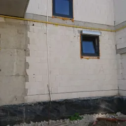 Surowy stan budynku w trakcie budowy: widoczne okna z roletami, żółte rury gazowe, zabezpieczenie fundamentów folią, tymczasowe drzwi wejściowe.