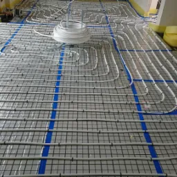 Rozłożona instalacja ogrzewania podłogowego na folii izolacyjnej z siatką, widoczny zwój rur i taśma montażowa.