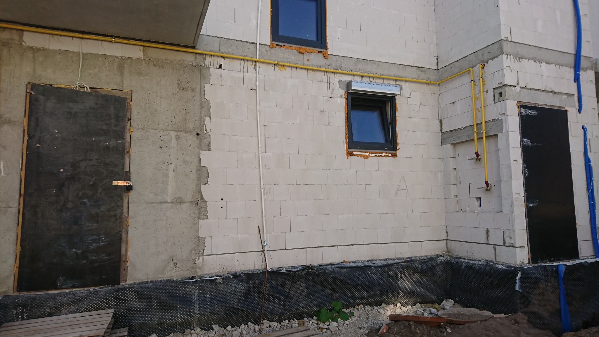 Surowy stan budynku w trakcie budowy: widoczne okna z roletami, żółte rury gazowe, zabezpieczenie fundamentów folią, tymczasowe drzwi wejściowe.
