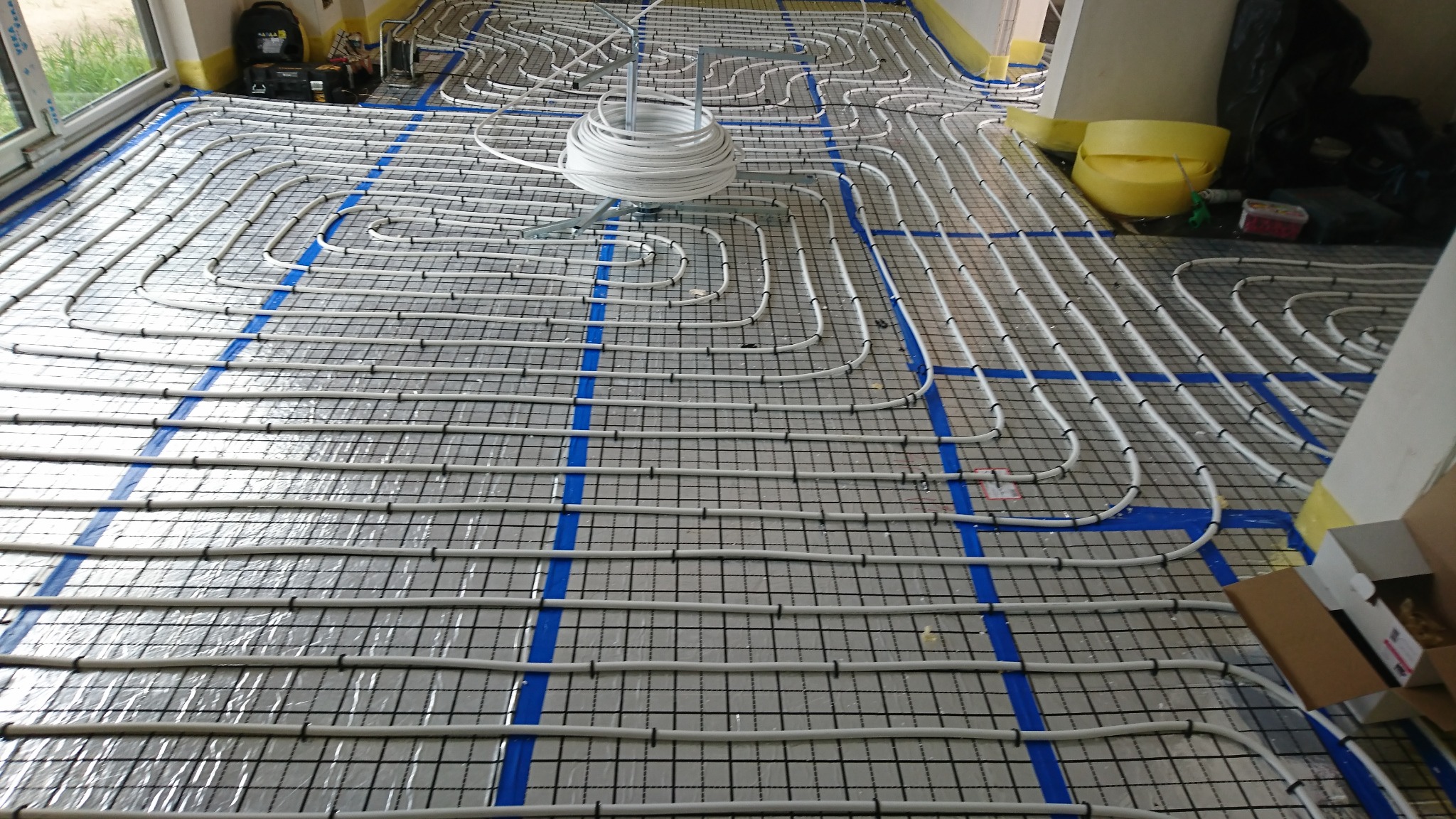 Rozłożona instalacja ogrzewania podłogowego na folii izolacyjnej z siatką, widoczny zwój rur i taśma montażowa.
