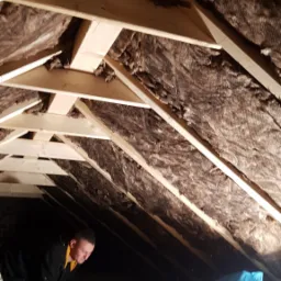 Prace izolacyjne na poddaszu: widoczne drewniane krokwie oraz warstwa izolacji z folią, pracownik w czarnej bluzie pochyla się nad podłogą, obok leży niebieski worek.