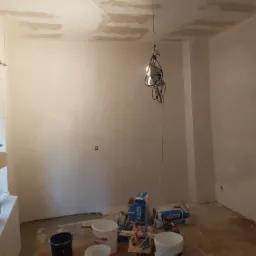Wnętrze pokoju w trakcie prac wykończeniowych, widoczne ślady gładzi na suficie, worki z zaprawą, wiadra i narzędzia na podłodze, wisząca lampa robocza.