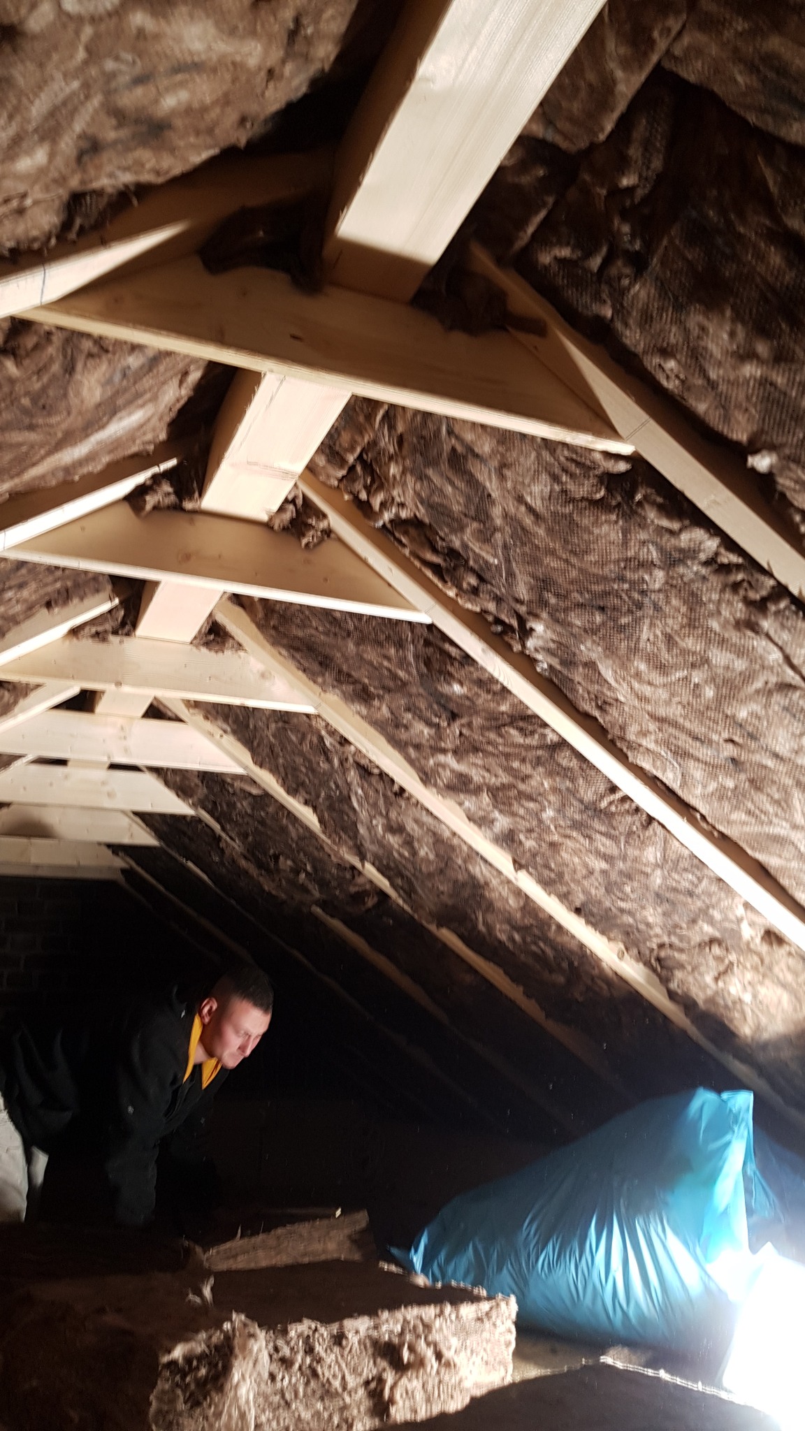 Prace izolacyjne na poddaszu: widoczne drewniane krokwie oraz warstwa izolacji z folią, pracownik w czarnej bluzie pochyla się nad podłogą, obok leży niebieski worek.