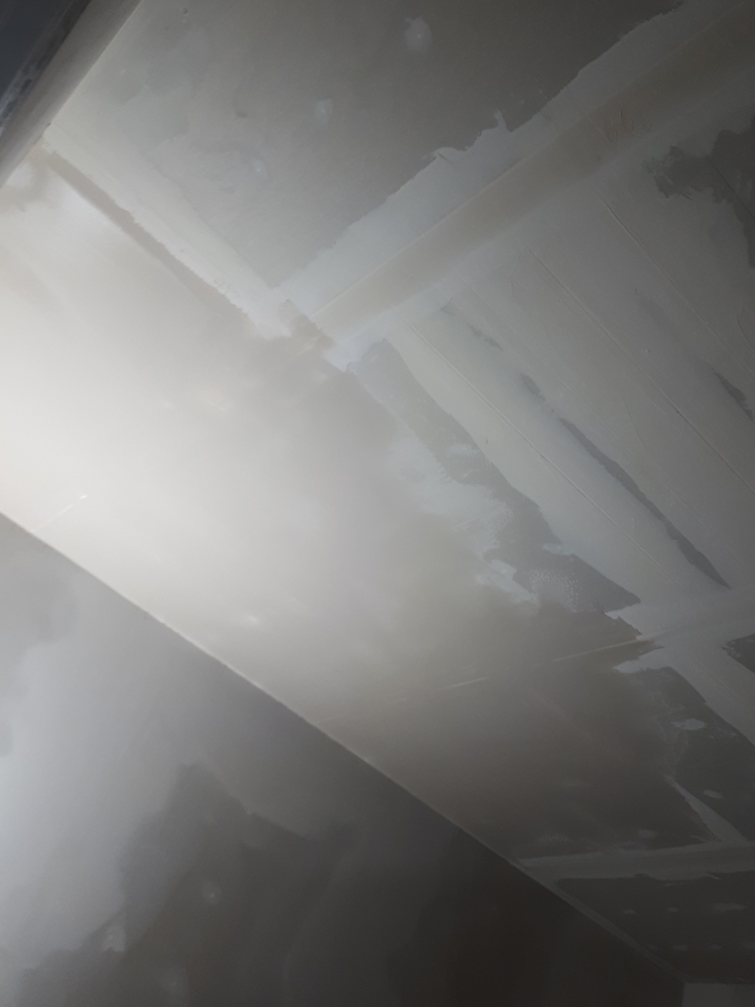 Niedokończona powierzchnia sufitu z widocznymi pasami świeżo nałożonej gładzi szpachlowej, nierówności i niedoskonałości w oświetleniu z boku.