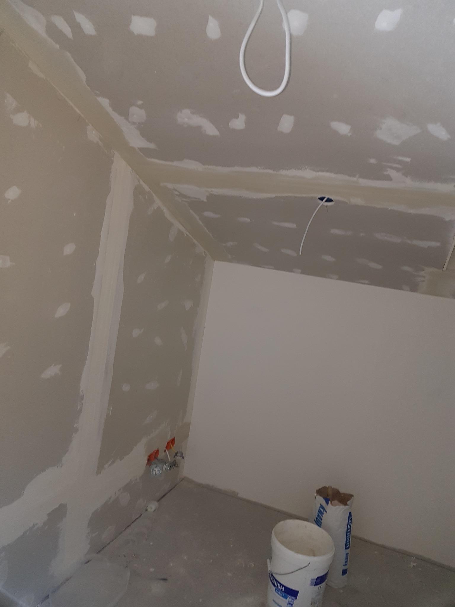 Wnętrze w trakcie prac wykończeniowych: ściany i sufit pokryte płytami gipsowo-kartonowymi, widoczne łączenia zaszpachlowane, puszki elektryczne wystające ze ściany oraz wiadro i worek z materiałem...