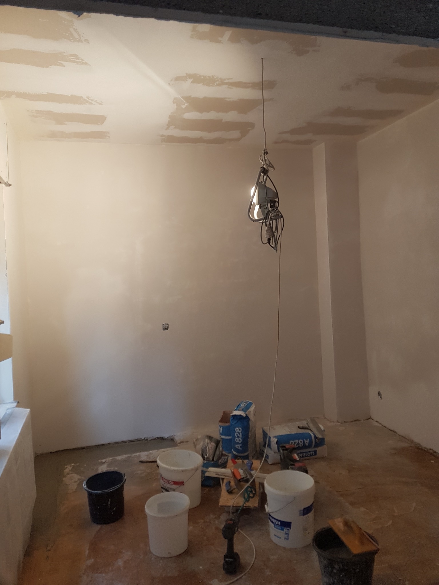 Wnętrze pokoju w trakcie prac wykończeniowych, widoczne ślady gładzi na suficie, worki z zaprawą, wiadra i narzędzia na podłodze, wisząca lampa robocza.