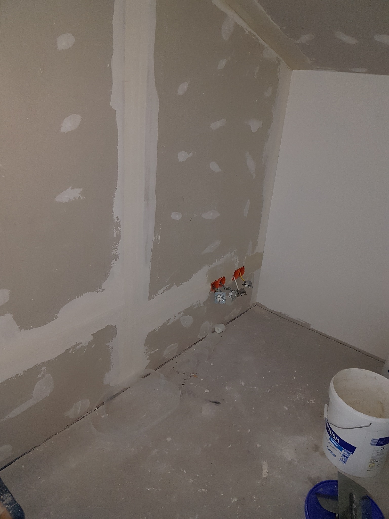 Przygotowanie ścian do wykończenia: płyty gipsowo-kartonowe z widocznymi łączeniami i punktowymi szpachlowaniami, puszki elektryczne wystające ze ściany, surowe podłoże podłogi, wiadro z masą...