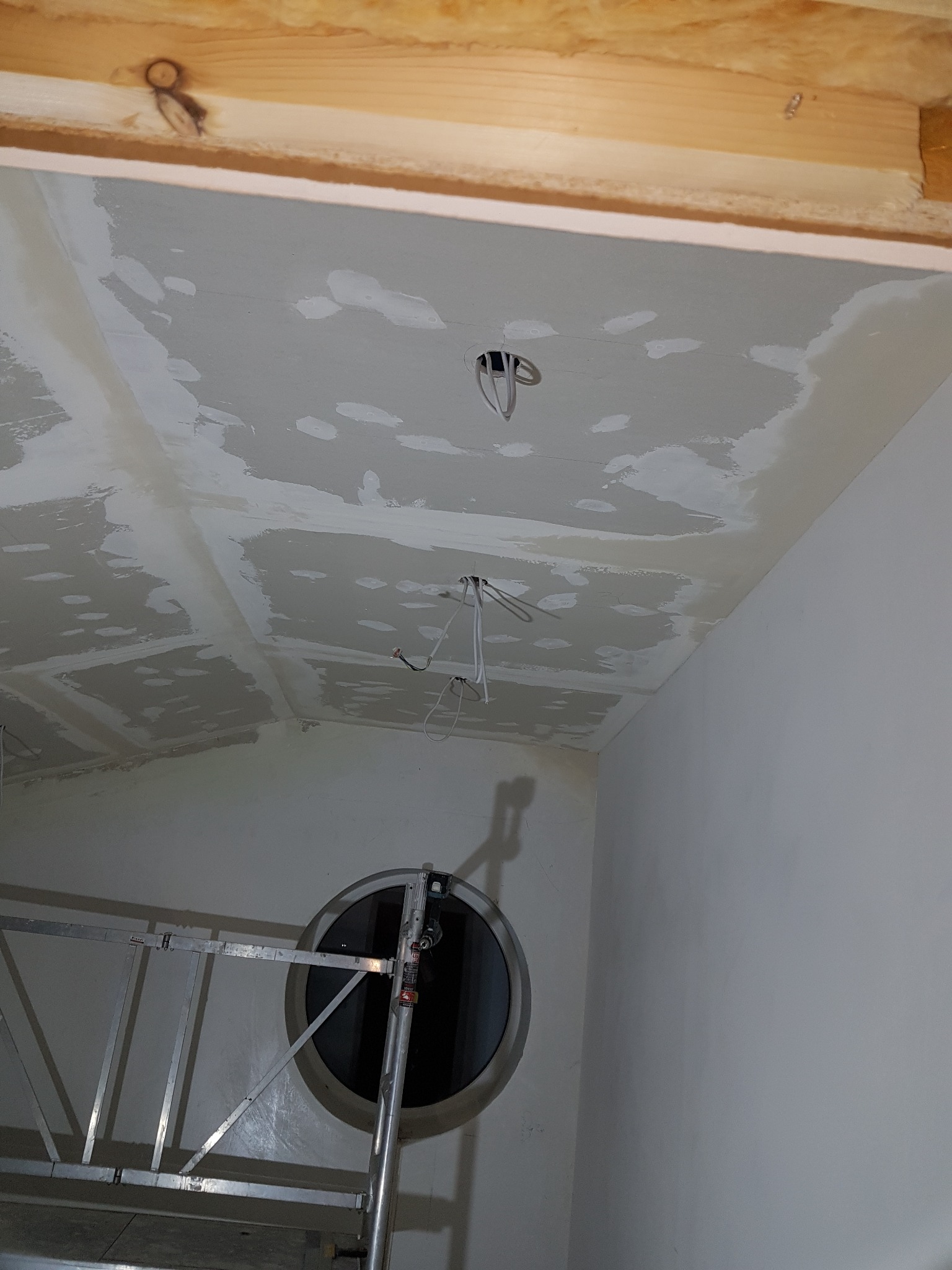 Poddasze w trakcie prac wykończeniowych: widoczne płyty kartonowo-gipsowe z nałożoną, nierównomierną warstwą szpachli, wystające przewody elektryczne i aluminiowy podest roboczy z okrągłym oknem...