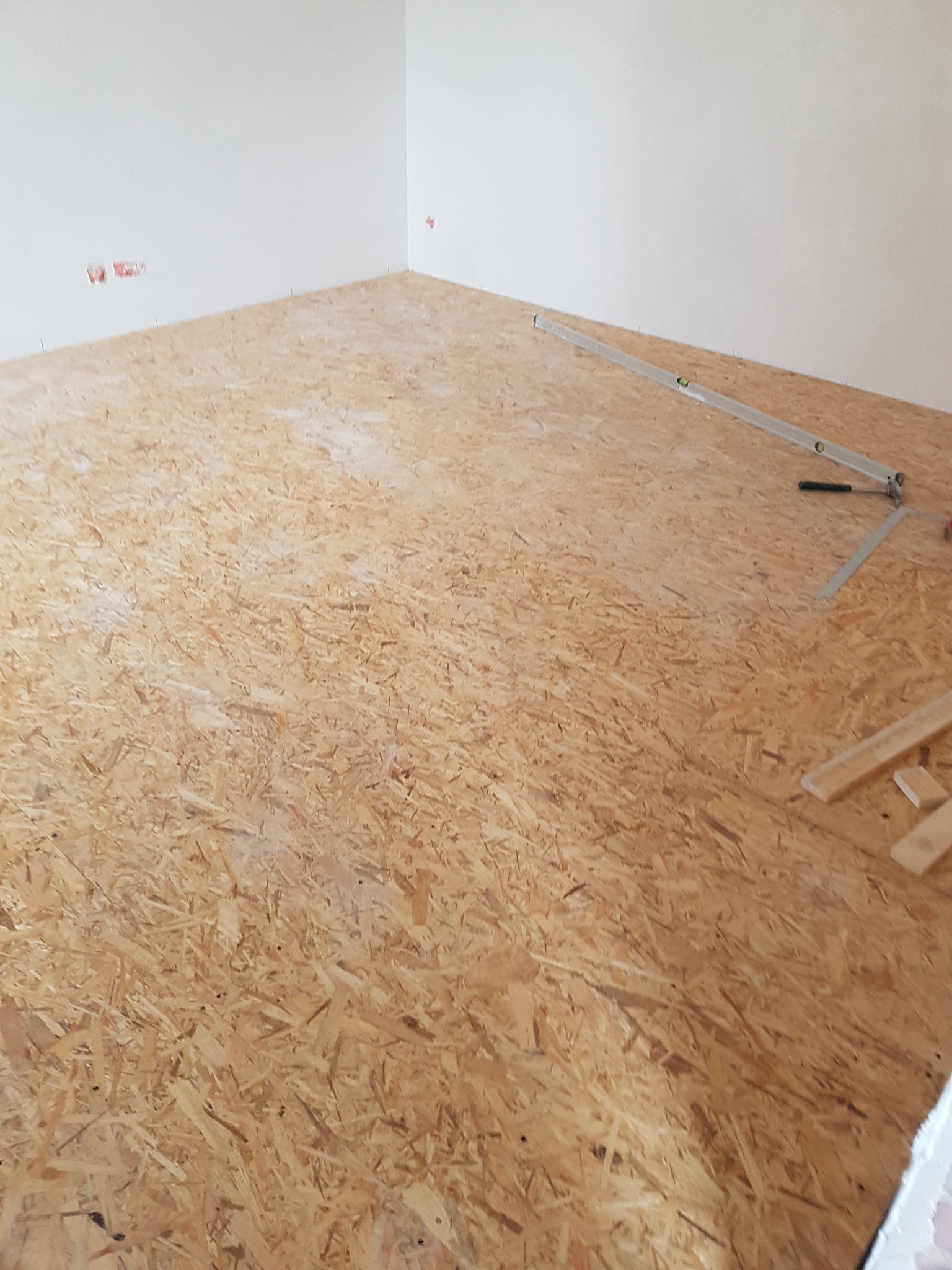 Płyty OSB ułożone na podłodze w pustym pomieszczeniu, poziomica i fragmenty drewnianych listew na powierzchni.