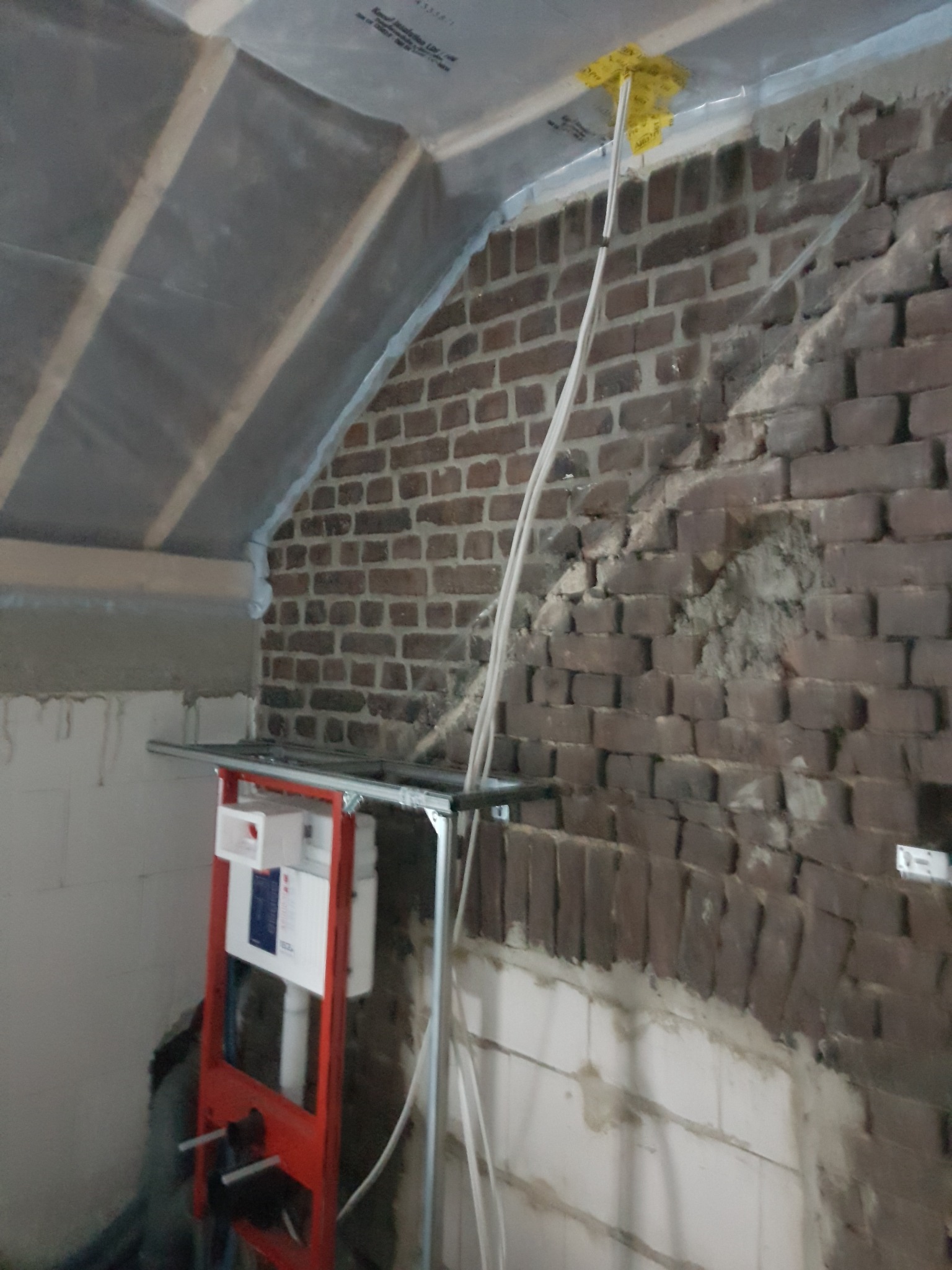 Poddasze w trakcie prac wykończeniowych: widoczna ceglana ściana z częściowo uzupełnionymi ubytkami, zamontowany stelaż podtynkowy WC na tle surowej ściany, folia paroizolacyjna na skośnym suficie.