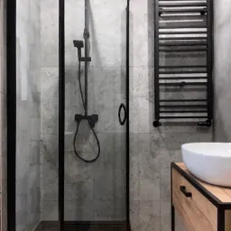 Nowoczesna łazienka z kabiną prysznicową typu walk-in z czarnymi profilami, grzejnikiem drabinkowym i umywalką nablatową. Szare płytki imitujące beton.