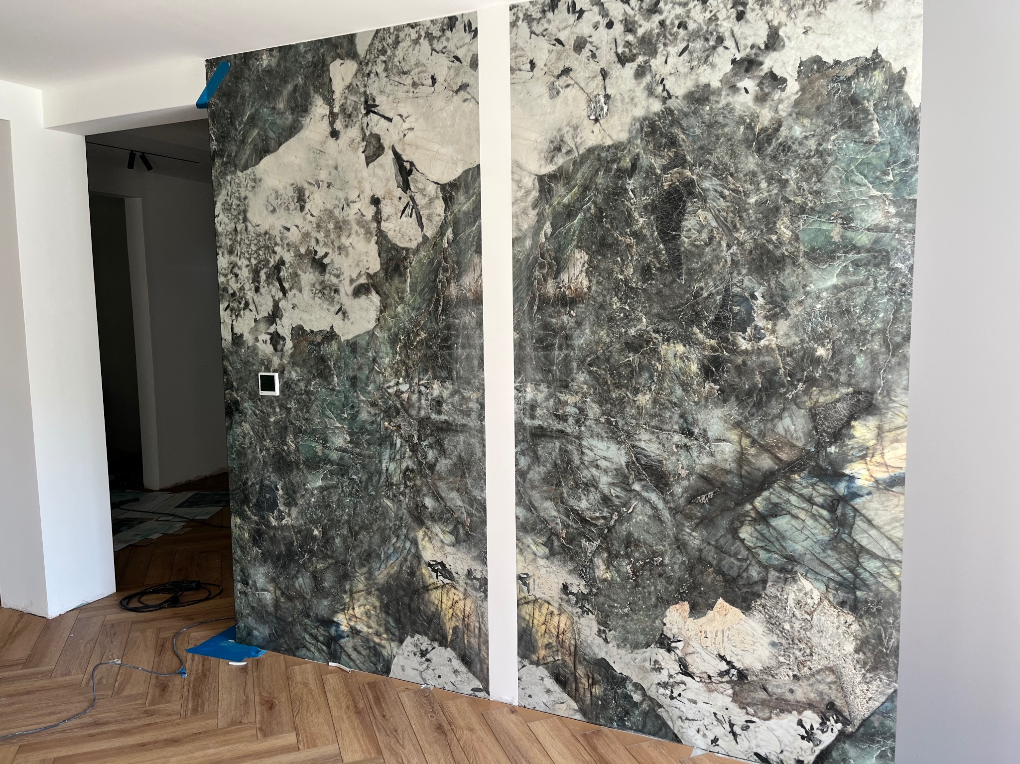 Montaż tapety imitującej marmur na ścianie w trakcie wykańczania wnętrza. Widoczne fragmenty podłogi w jodełkę i przygotowania do dalszych prac.