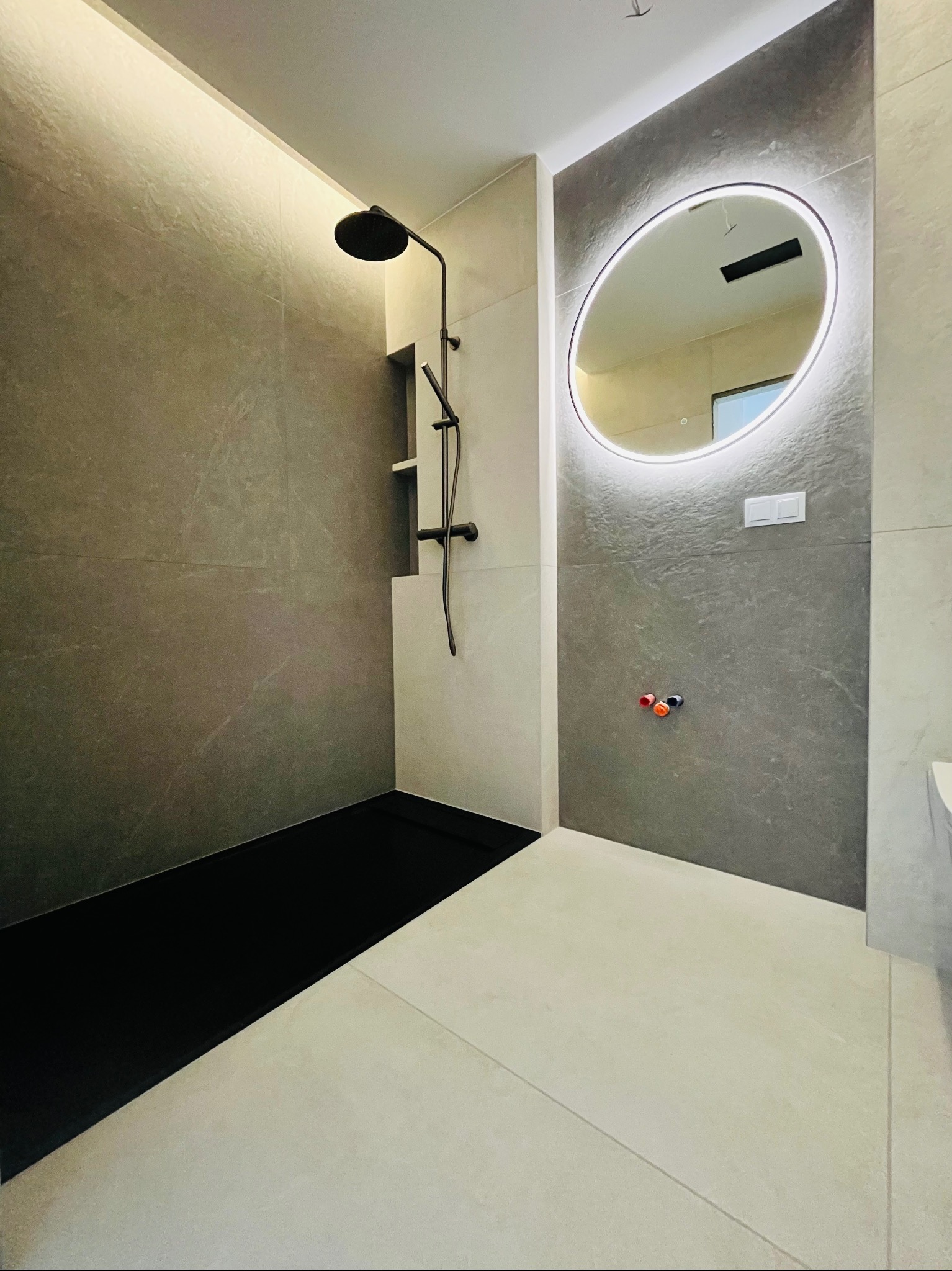 Nowoczesna łazienka z czarnym brodzikiem, szarymi płytkami i okrągłym lustrem z podświetleniem LED. Minimalistyczny design, widoczne przyłącza wodne.