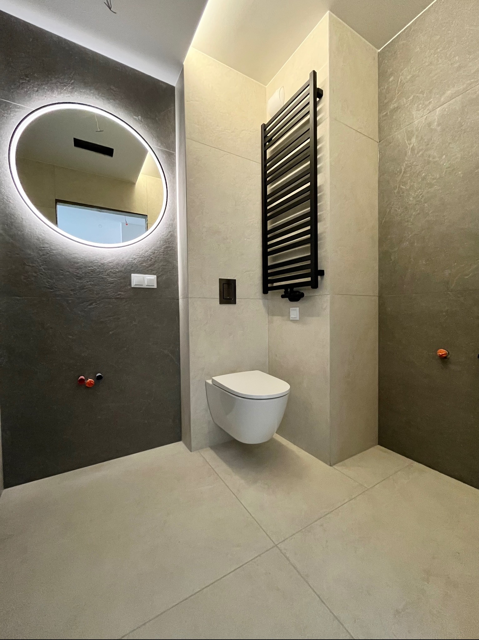 Nowoczesna łazienka po remoncie: wisząca toaleta, grzejnik drabinkowy, okrągłe lustro z podświetleniem LED i płytki w odcieniach szarości i beżu. Minimalistyczny design.