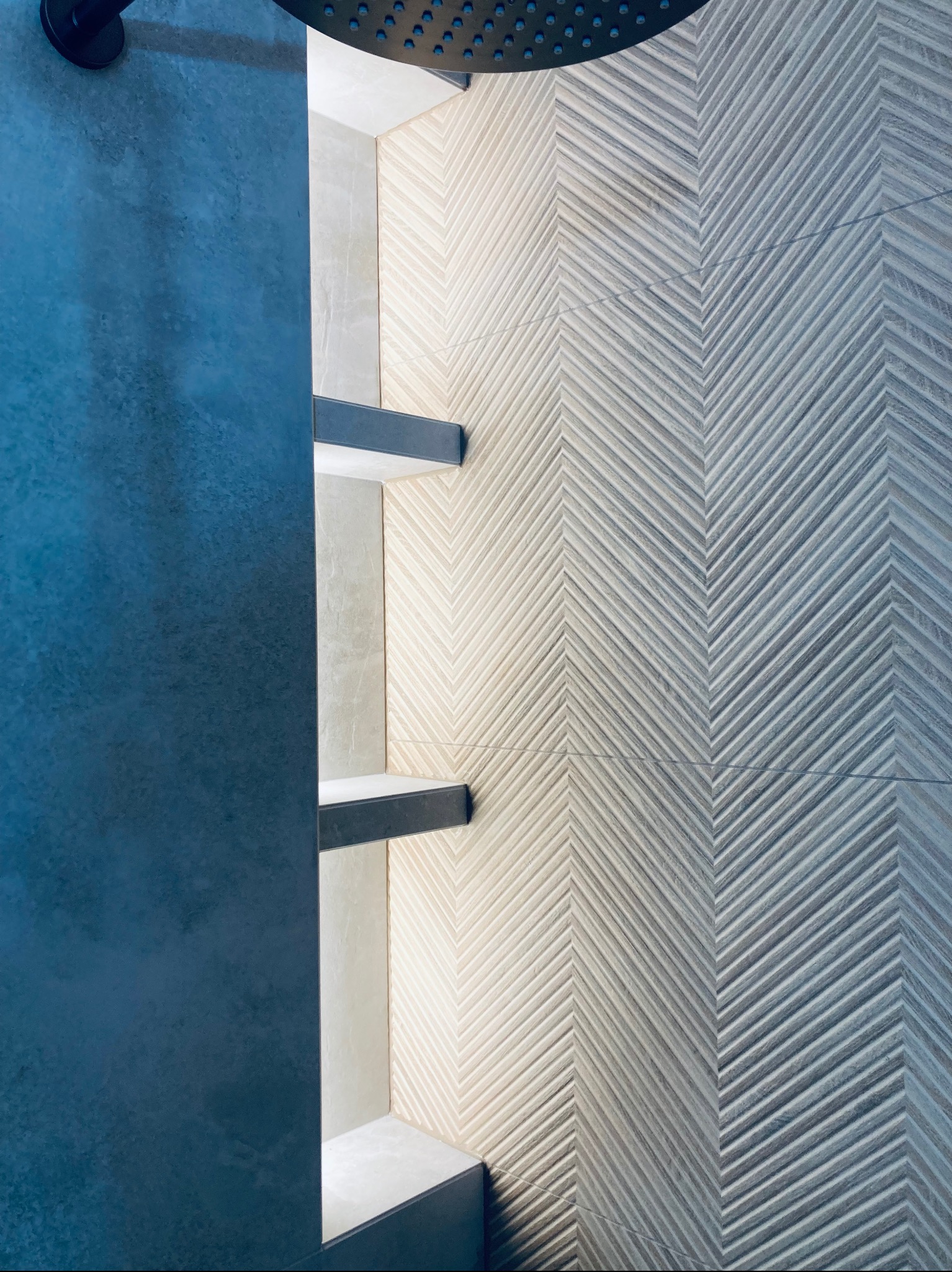 Narożnik prysznicowy z geometrycznym wzorem płytek w jodełkę i wbudowanymi półkami. Nowoczesny design z ciemną armaturą i niebieskim panelem bocznym.