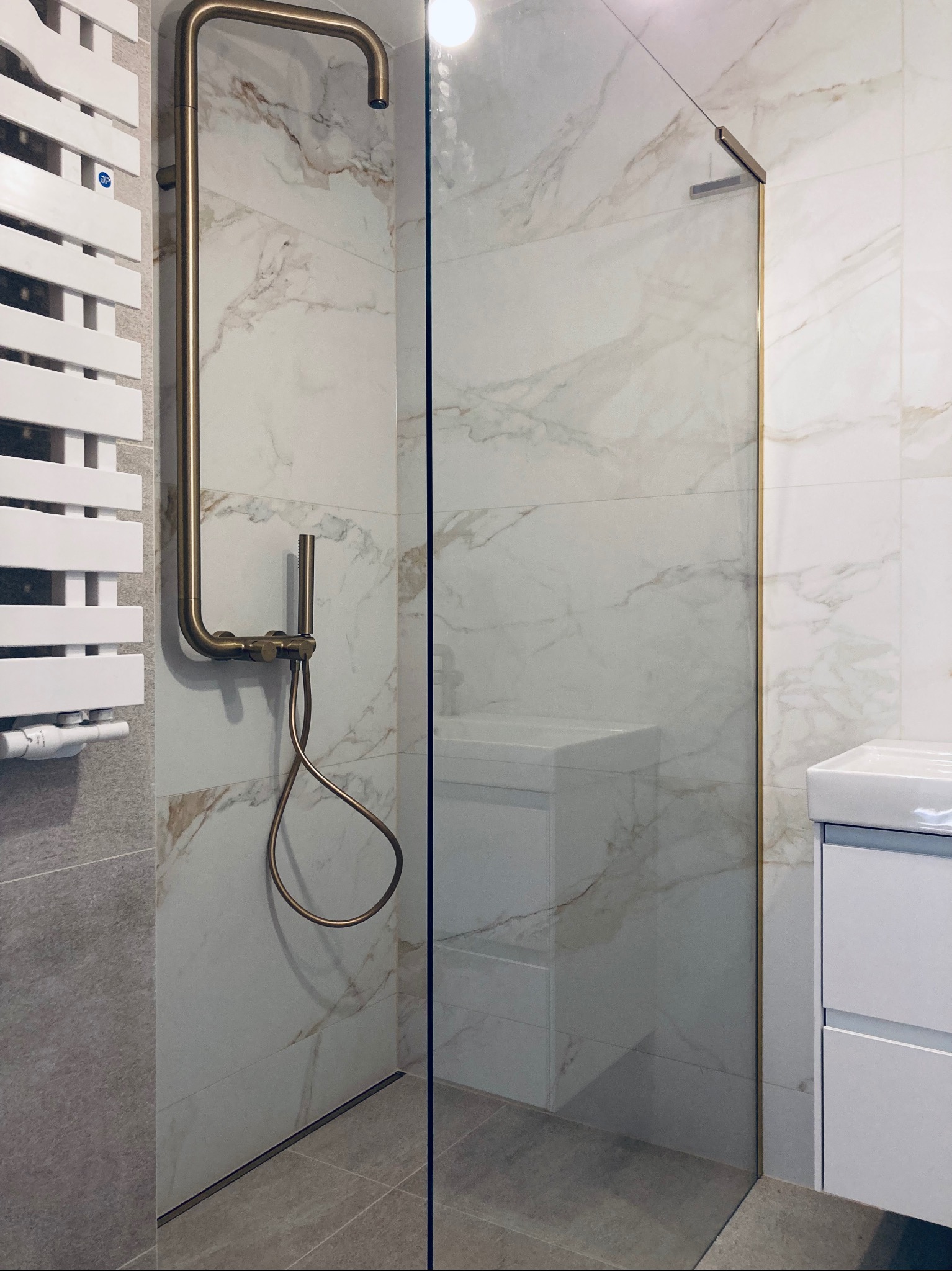 Nowoczesna kabina prysznicowa ze złotymi detalami: armatura, rama szklana, białe płytki imitujące marmur, grzejnik drabinkowy, umywalka z szafką w tle.