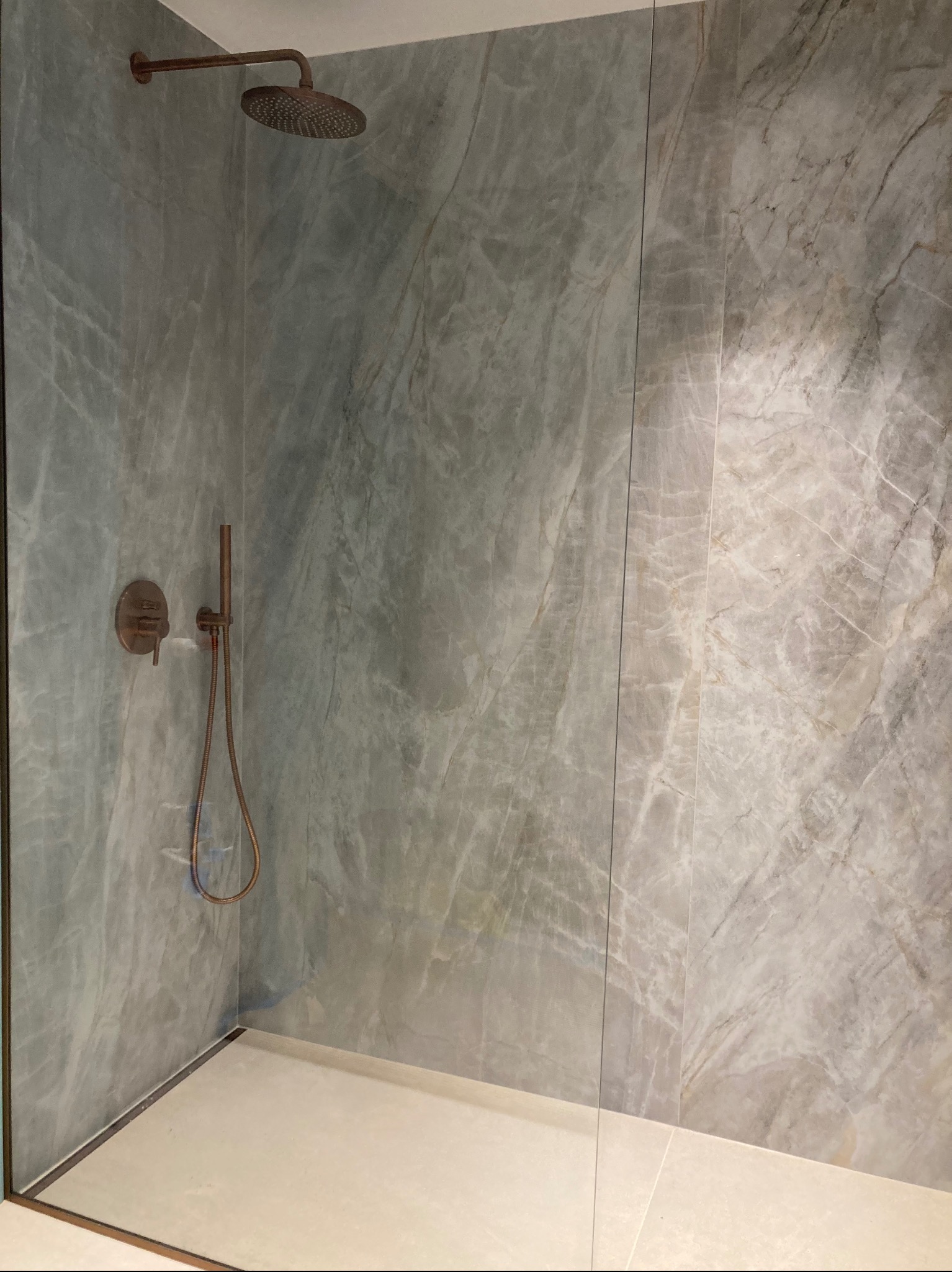 Nowoczesna kabina prysznicowa z marmurowymi płytkami w odcieniach szarości i beżu, wyposażona w miedzianą armaturę i szklaną ściankę. Minimalistyczny design.