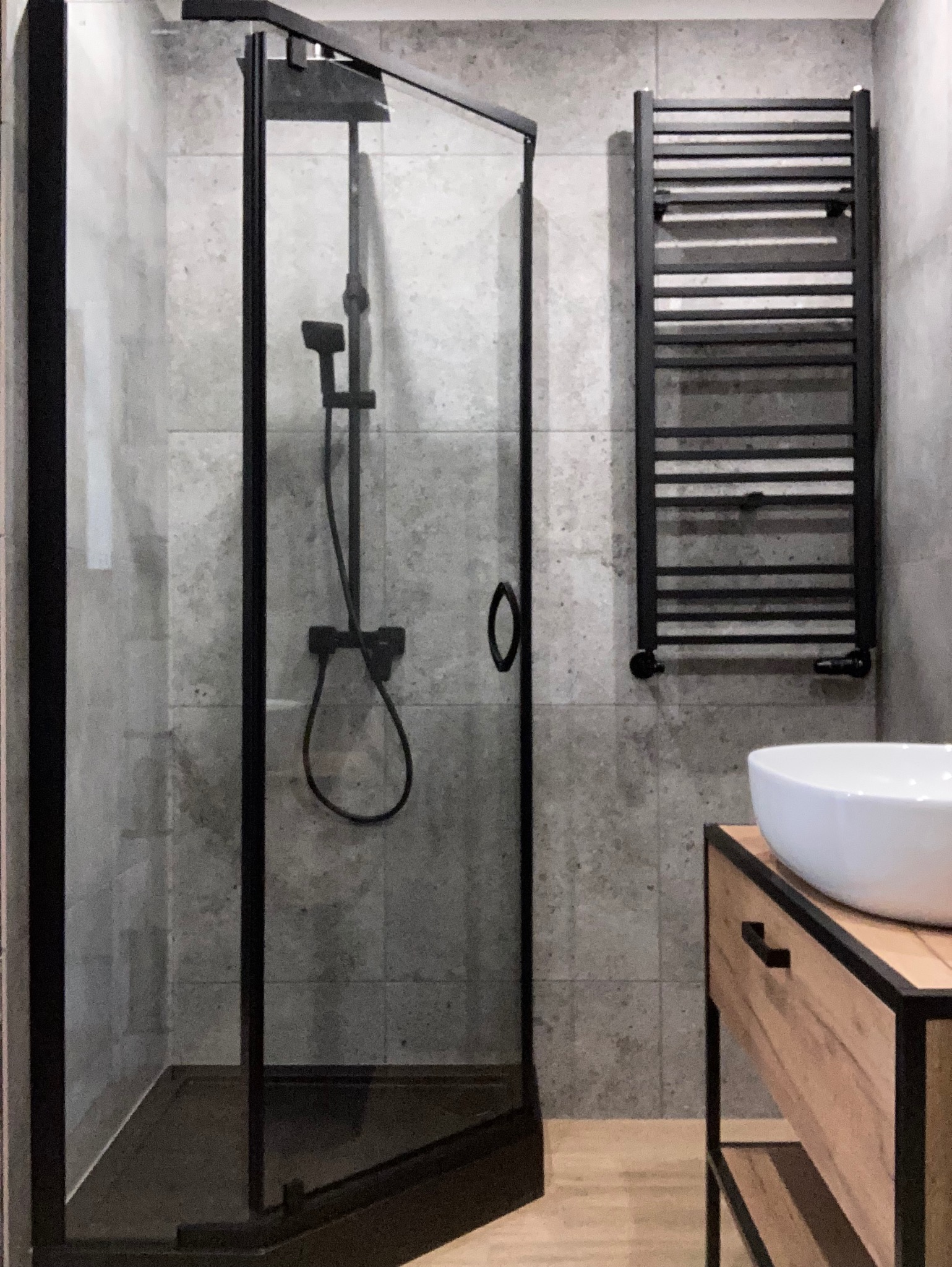 Nowoczesna łazienka z kabiną prysznicową typu walk-in z czarnymi profilami, grzejnikiem drabinkowym i umywalką nablatową. Szare płytki imitujące beton.