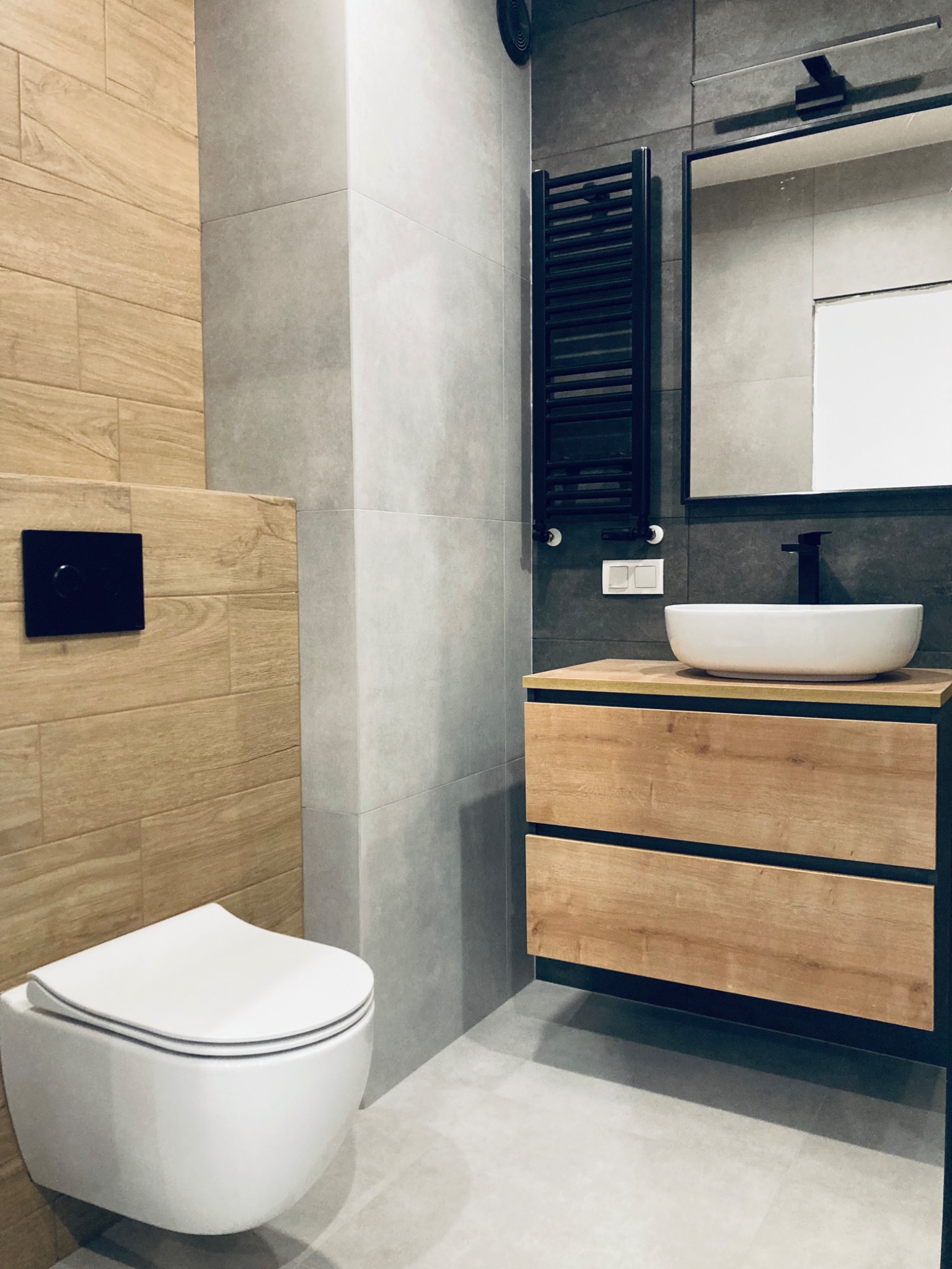 Nowoczesna łazienka z drewnianymi akcentami: wisząca toaleta, umywalka na szafce z szufladami, grzejnik drabinkowy i szare płytki. Minimalistyczny design.