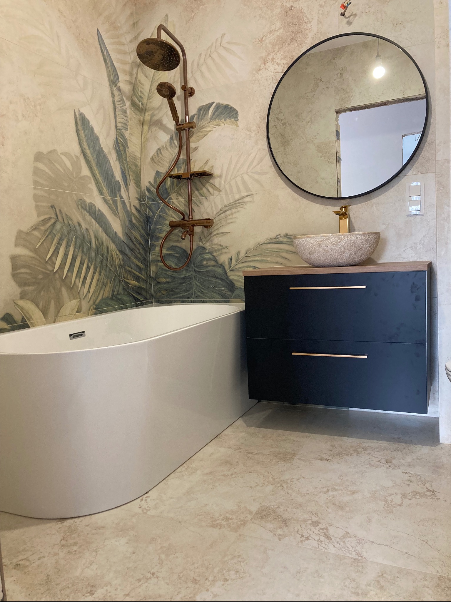 Nowoczesna łazienka z wanną, okrągłym lustrem, umywalką na szafce w kolorze granatowym i baterią w kolorze złotym. Na ścianie płytki z motywem roślinnym.